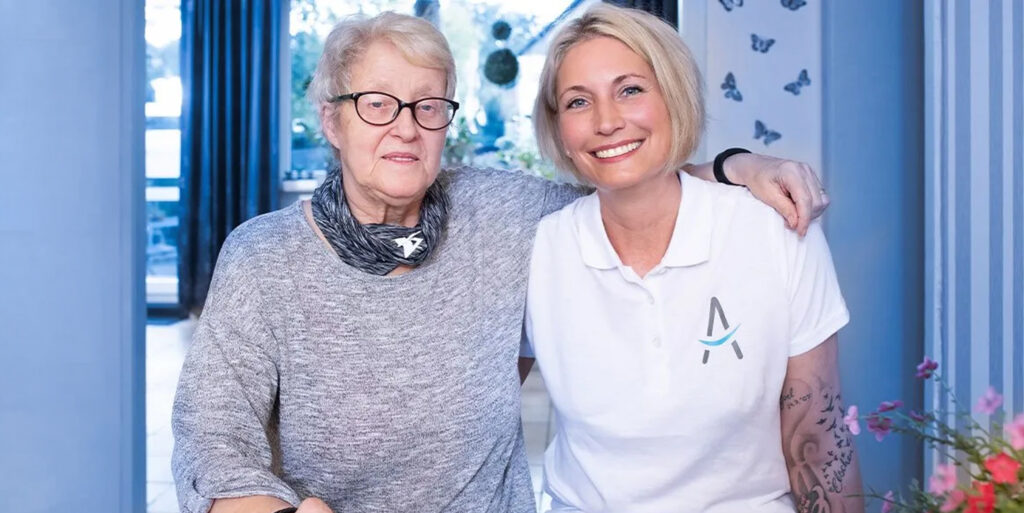 Pflegekraft in weißem Poloshirt sitzt arm in Arm mit einer Seniorin in deren Zuhause und begleitet sie freundlich im Alltag.